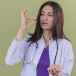 young woman doctor wearing white coat and with stethoscope holding pill with fingers looking at it with serious face standing over green background